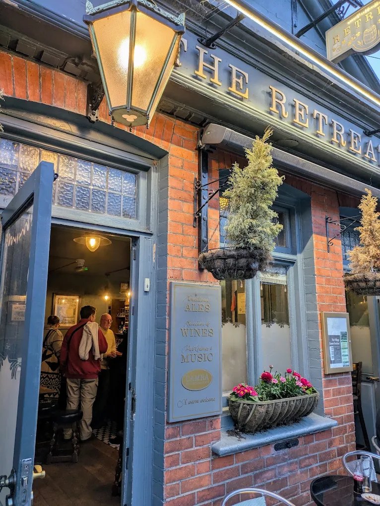 The Retreat at dusk: the lantern lit, the door open onto regulars at the bar, gold lettering on slate-blue paintwork and a window box of pink flowers